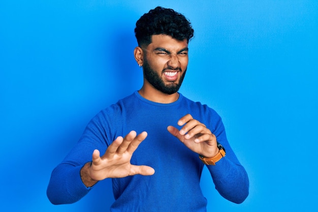 Arab man with beard wearing casual blue sweater disgusted expression displeased and fearful doing disgust face because aversion reaction with hands raised