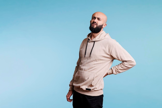 Arab man with lower back injury and looking at camera with painful facial expression. Grimacing young person suffering from backache and holding hand on sore muscles portrait