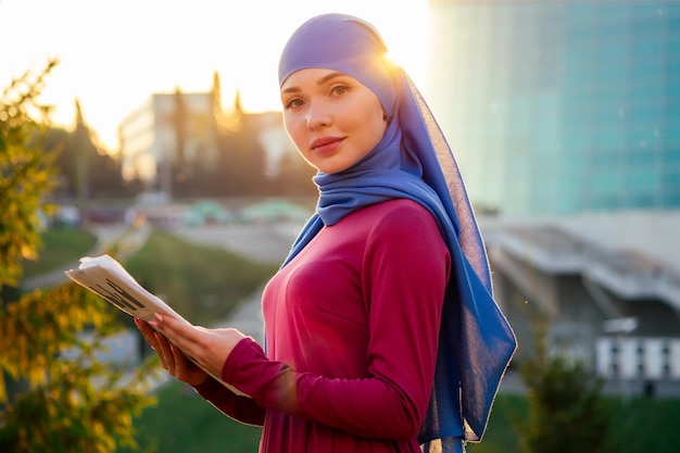 Arab Muslim business woman hijab with makeup standing on the street holding documents and papers on a background of summer park woman is dressed in a stylish abaya and shawl successful deal concept