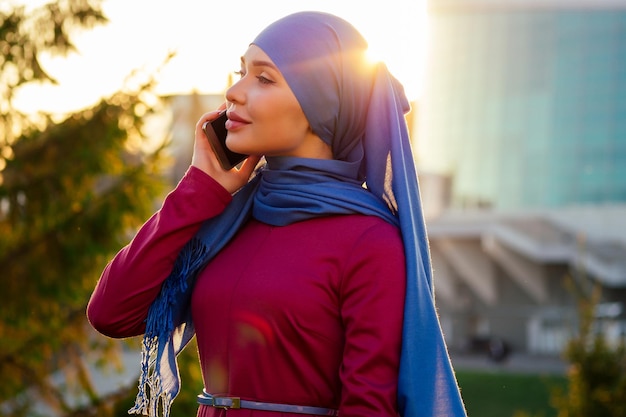 Arab Muslim business woman hijab with makeup standing on the street talking on the phone on a background of summer park The woman is dressed in a stylish abaya and shawl