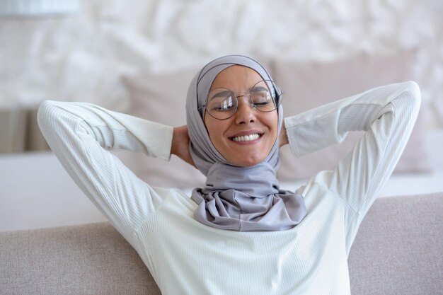 Arab young female student in hijab and glasses sitting on sofa at home hands behind head eyes closed