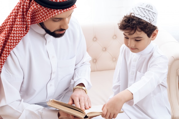 Photo arabian bearded man is reading book with small son.