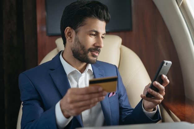 Photo arabian businessman holding credit card using  mobile phone shopping online sitting in airplane