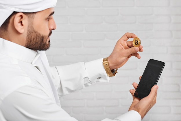 黄金のbitcoinと携帯電話を維持する伝統的な白のイスラム教徒のアラビアのビジネスマン