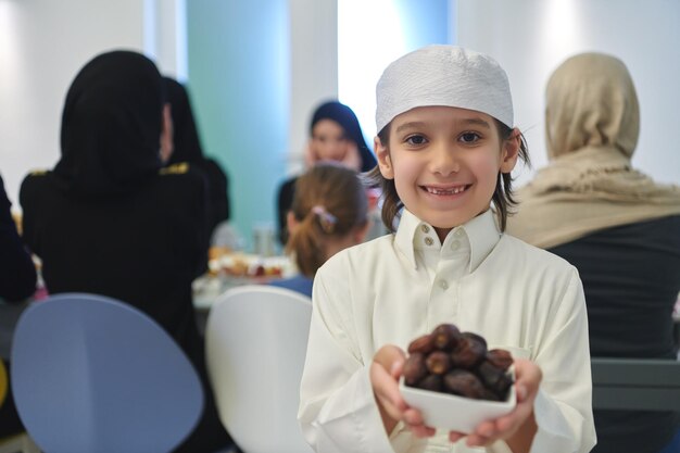 Photo arabian kid in the traditional clothes during iftar. portrait of happy young muslim boy holding bowl of dates while rest of the family are eating