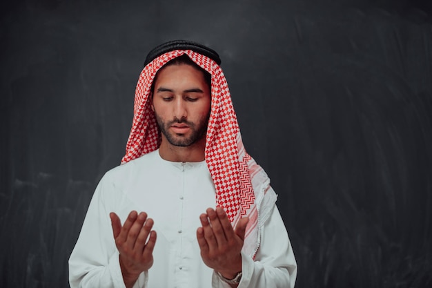 Arabian man in traditional clothes making traditional prayer to God, keeps hands in praying gesture in front of black chalkboard representing modern islam fashion and ramadan kareem concept.