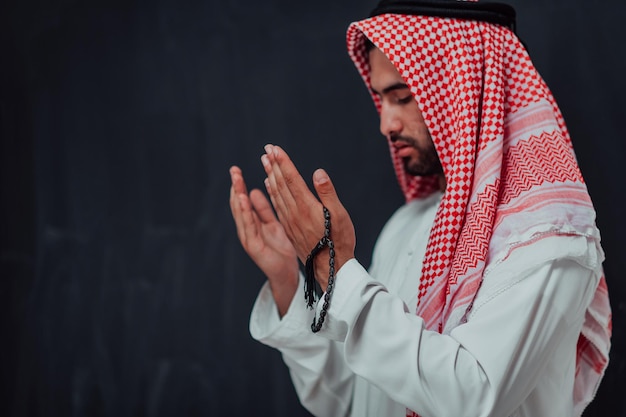 Photo arabian man in traditional clothes making traditional prayer to god, keeps hands in praying gesture in front of black chalkboard representing modern islam fashion and ramadan kareem concept.