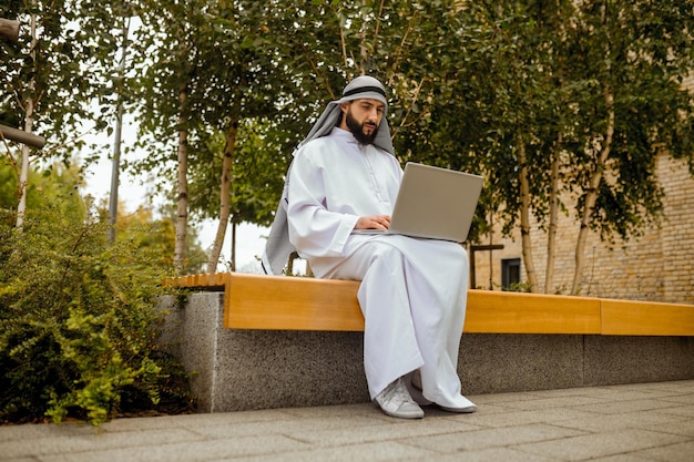 An arabian man in traditional clothing working on a laptop outside