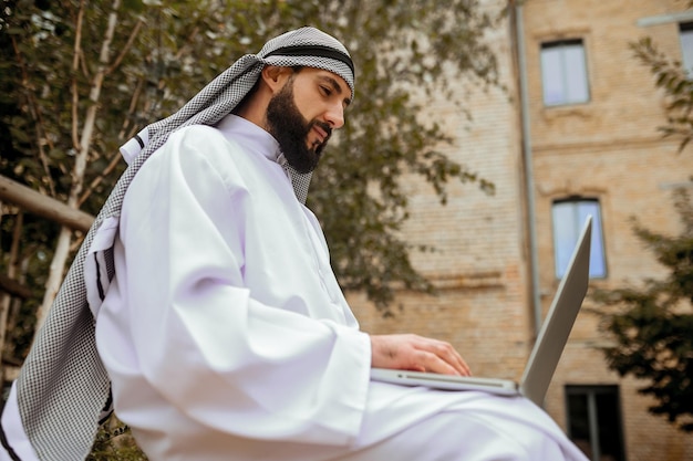 An arabian man in traditional clothing working on a laptop outside