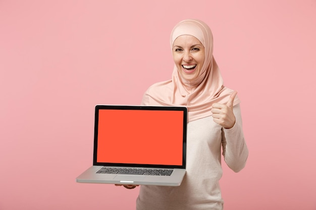 Arabian muslim girl in hijab light clothes posing isolated on pink background. People religious lifestyle concept. Mock up copy space. Hold laptop pc computer with blank empty screen showing thumb up.