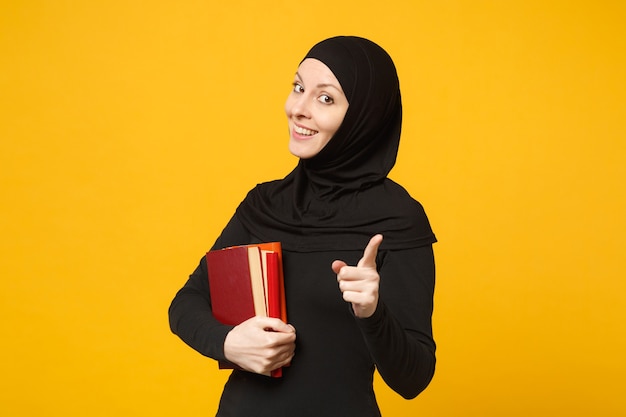 ヒジャーブの黒い服を着たアラビアのイスラム教徒の学生の女の子は、黄色の壁の肖像画に分離された本を保持しています。人々の宗教的なライフスタイル、高校の概念の教育。 。