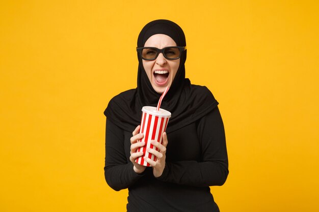 Hijab 검은 옷에 아라비아 무슬림 여성 3d imax 안경 시계 영화 필름 노란색 벽 초상화에 고립 된 소 다의 종이 컵을 개최. 사람들의 라이프 스타일 개념. .