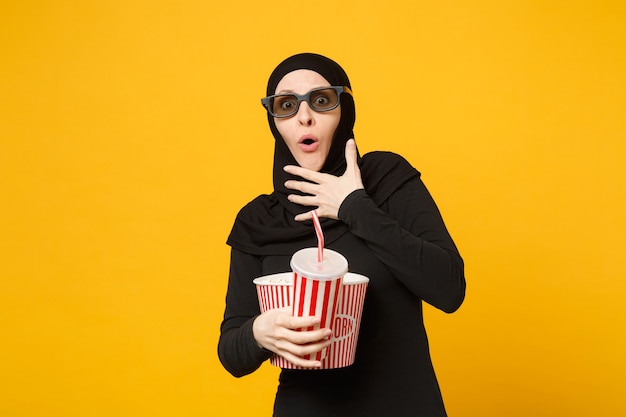 Arabian muslim woman in hijab black clothes 3d imax glasses watch movie film hold popcorn, cup of soda isolated on yellow wall portrait. people lifestyle concept