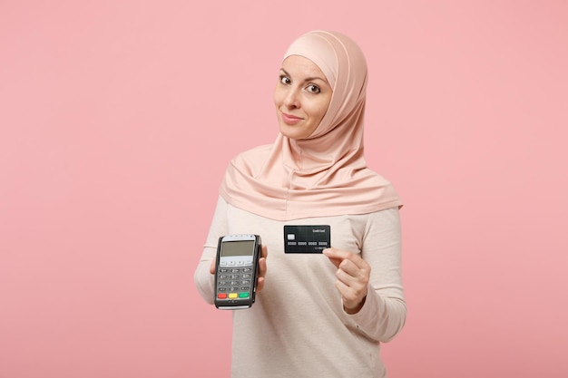 Arabian muslim woman in hijab light clothes posing isolated on pink background. People religious lifestyle concept. Mock up copy space. Hold payment terminal to process, acquire credit card payments.