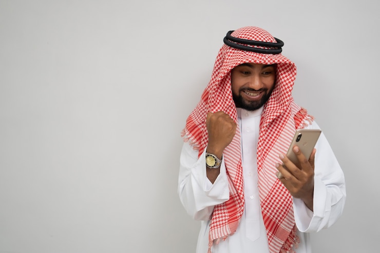 Photo an arabian young man in a turban using a mobile phone happily looking at the screen with an excited ...