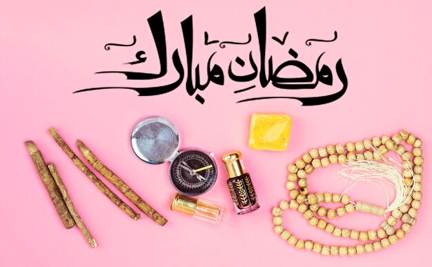 Arabic calligraphy meaning happy ramadan in english rosary siwak and perfume on pink background