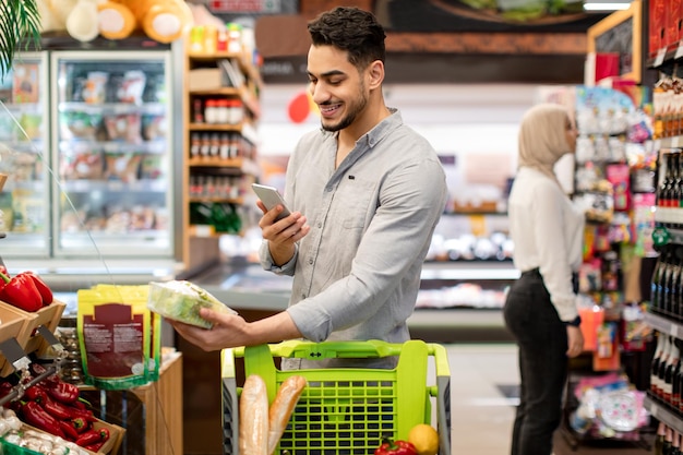 Arabic male using smartphone during shopping scanning vegetables in supermarket