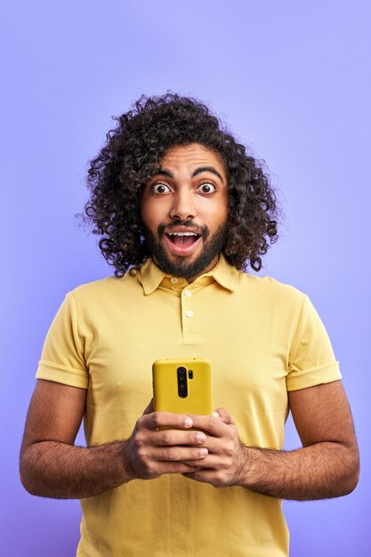 Arabic man being surprised after reading message on smartphone, curly male stand emotionally react on message, looking at camera with opened mouth