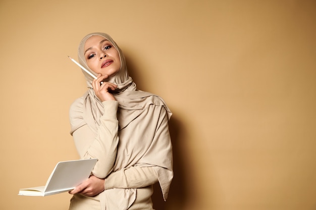 Arabic Muslim woman wearing beige hijab holds a diary and looks up thoughtfully, pointing with a white pencil on a beige with copy space.