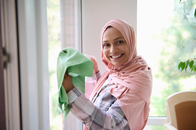 Arabic Muslim woman with head covered in pink hijab, smiles looking at camera, enjoying spring cleaning in the house on a sunny day, removing stains, washing windows in the veranda