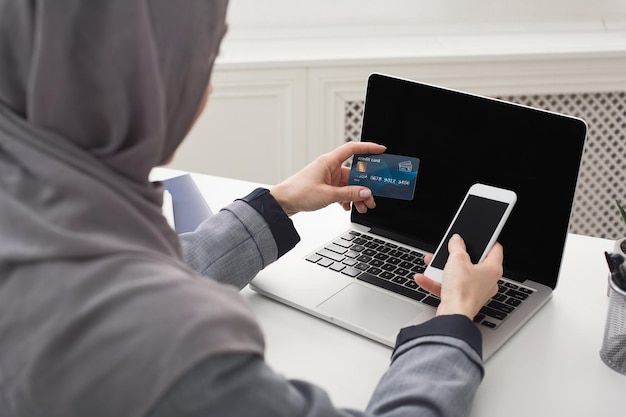 Arabic woman shopping online on laptop with credit card, back view, empty screens