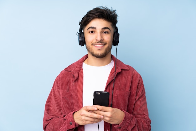 Arabische knappe mens op blauwe muur het luisteren muziek met mobiel en het kijken voorzijde