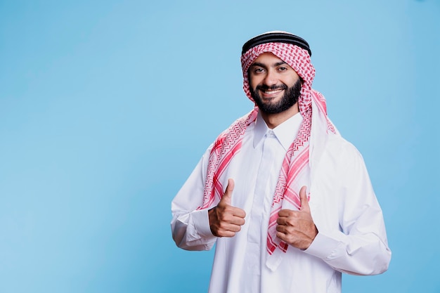 Arabische man die islamitische kleding draagt en duimen opgeeft en een vrolijk expressieportret toont. Lachende moslim persoon gekleed in traditioneel gewaad en hoofddoek en camera kijken