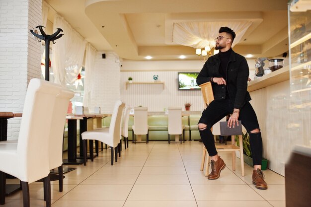 Arabische man draagt een zwarte spijkerjas en een bril in café drinkt koffie aan de bar met boek Stijlvolle en modieuze Arabische modelman