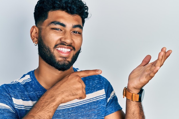 Arabische man met baard in casual gestreept t-shirt verbaasd en glimlachend naar de camera terwijl hij met de hand presenteert en met de vinger wijst.
