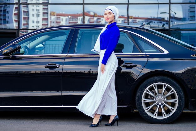 Arabische moslim zakenvrouw hijab met make-up staande voor haar luxe auto op straat op een achtergrond van wolkenkrabbers. De vrouw is gekleed in een stijlvolle abaya