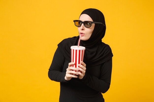 Arabische moslimvrouw in hijab zwarte kleding 3d imax bril kijken film film houd papieren kopje frisdrank geïsoleerd op gele muur portret. mensen levensstijl concept. .