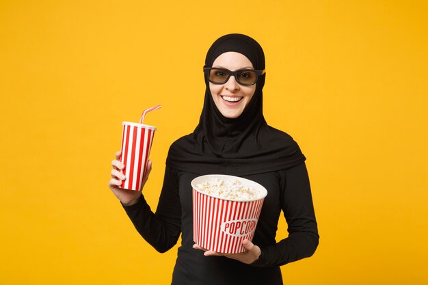 Arabische moslimvrouw in hijab zwarte kleding 3d imax bril kijken film film houd popcorn, kopje frisdrank geïsoleerd op gele muur portret. Mensen levensstijl concept. .