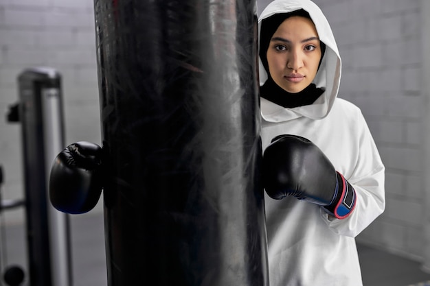 Arabische vrouwelijke bokser poseren in sportschool, jonge vrouw treinen met zware bokszak. portret van zelfverzekerd vrouwelijk werkuitje, een pauze nemen, in witte sportieve hijab