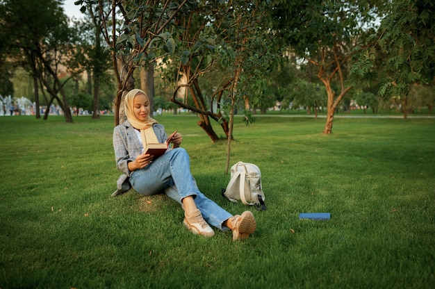 Arabische vrouwelijke student in hijab leerboek lezen in zomer park. Moslimvrouw met boeken die op het gazon rusten.