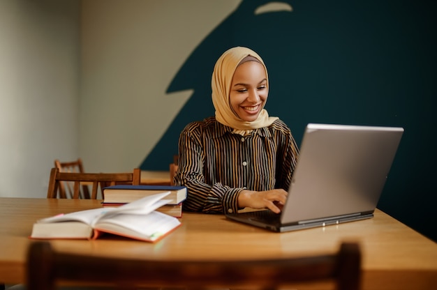 Arabische vrouwelijke student in hijab met laptop in het universiteitscafé. Moslimvrouw met boeken die in bibliotheek zitten.