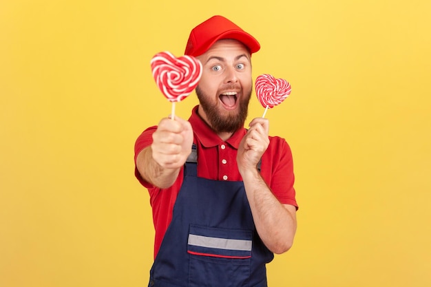 Arbeidersmens die blauw uniform draagt dat smakelijke suikerachtige lolly in de vorm van een hart vasthoudt met een opgewonden uitdrukking