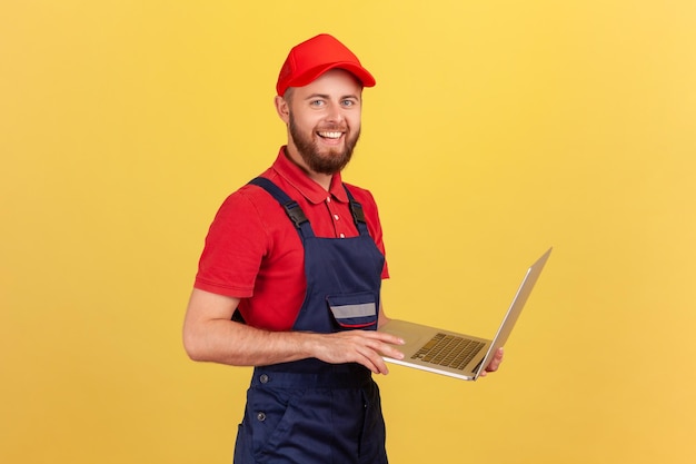 Arbeidersmens die blauwe overall rode t-shirt en pet dragen die aan laptop werken die positieve emoties uitdrukken