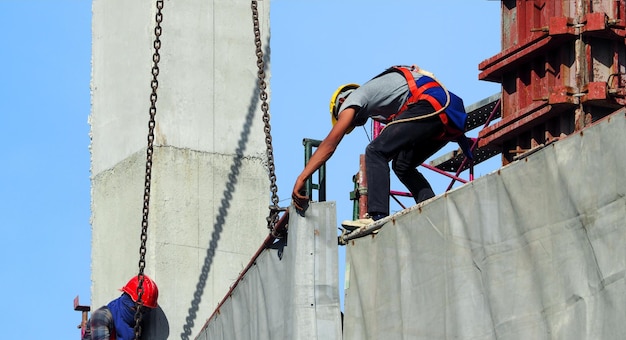 Arbeidsmens die aan bouwwerf met helm en veiligheidsuitrusting werkt en het gebouw bedekt met grijze kleurenvinyl en blauwe lucht.