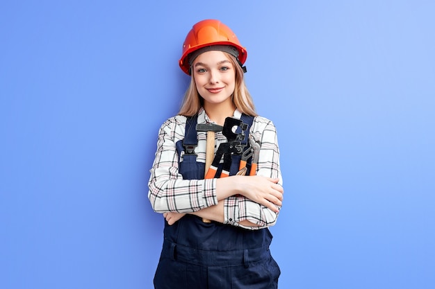 Architetto donna d'affari in casco arancione in piedi contro il muro blu, giovane femmina caucasica in tuta da lavoro uniforme come ingegnere costruttore