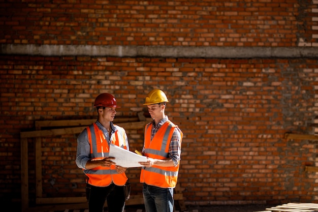 Архитектор и руководитель строительства, одетые в оранжевые рабочие жилетки и шлемы, обсуждают документацию на фоне кирпичной стены.
