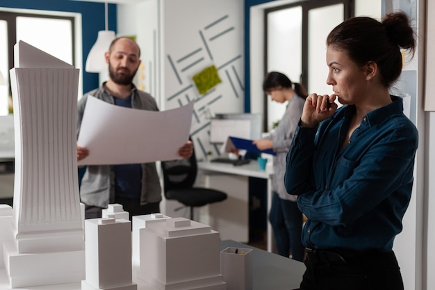 Architect die in de verte kijkt en naar het schaalmodel van de wolkenkrabber kijkt met een teamcollega-ingenieur die bouwplannen vasthoudt. Team van architecten die architecturale ideeas brainstormen.