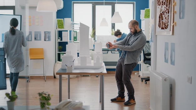 Architect die mobiele telefoon gebruikt om blauwdrukkenplan te maken met bouwmodel en maquette voor bouwontwikkeling. man ingenieur die met smartphone werkt om strategieproject te ontwerpen.