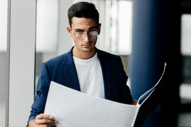 Architect dressed in stylish clothes stands next to window and explores the sheet with drawing in his hands .