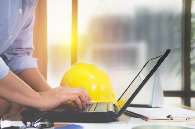 Architect engineer using laptop for working with yellow helmet on desk.