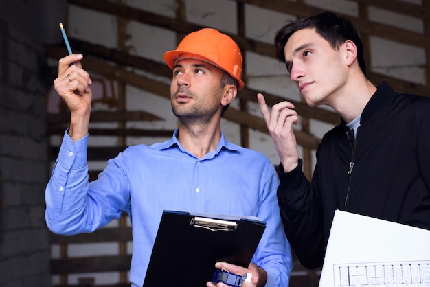 Ingegnere architetto che indossa un casco arancione sul luogo di lavoro che spiega i piani del progetto al cliente