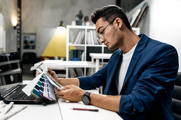 The architect in glasses and in a blue jacket is working with catalog of colors on the desk in the office .