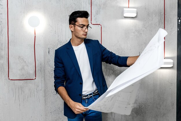 Architect in glasses dressed in blue checkered jacket and jeans works with blueprints on the background of the concrete wall .