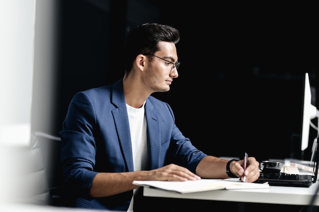 Architect in glasses dressed in blue checkered jacket makes notes in a notebook on the desk with laptop in the office .