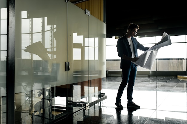 Architect in stijlvolle kleding houdt blad met tekening in zijn hand en praat via de telefoon op de achtergrond van een modern glazen gebouw met meerdere verdiepingen.