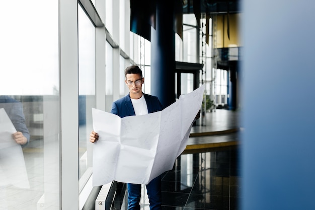 Architect in stijlvolle kleding houdt blad met tekening in zijn hand en praat via de telefoon op de achtergrond van een modern glazen gebouw met meerdere verdiepingen.